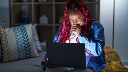 African American woman with braided hair using computer laptop at night feeling unwell and coughing as symptom for cold or bronchitis.